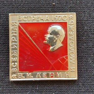 May include: A square gold-toned metal pin featuring a red enamel background with a raised gold profile portrait of Vladimir Lenin. The border contains the Cyrillic text "ВСЕМИНАРНАЯ ВСТРЕЧА 1970 И МОЛОДЕЖЬ" and "В.И. ЛЕНИН" embossed in gold lettering.
