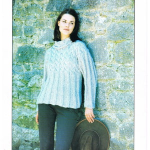 May include: A light blue knitted sweater with a cable knit pattern. The sweater has a high neck and long sleeves. The model is wearing black pants and a brown hat.