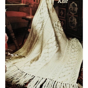 May include: A white knitted throw blanket with a cable knit pattern and fringe. The blanket is draped over a chair in a room with candles and a wooden cabinet. The text "Fisherman Knit" is printed on the image.