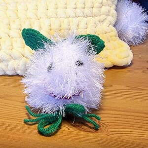 May include: A handmade white fuzzy creature with green ears and green tendrils. The creature has black eyes and a red mouth. It is lying on a yellow and white knitted blanket. The background is a wooden surface.
