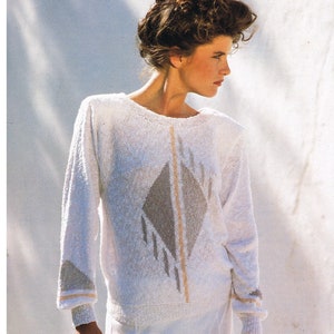 May include: A white sweater with a geometric design in shades of grey and beige. The sweater has a round neckline and long sleeves. The model is wearing white trousers. The text "HJERTE SILVER" is printed at the top of the image. The text "HJERTEGARN danish design" is printed at the bottom of the image.