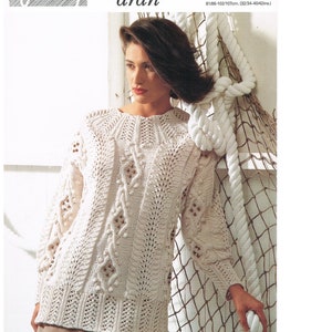 May include: A white aran knit sweater with a cable stitch pattern. The sweater is shown on a model against a background of a white rope and a brown net.