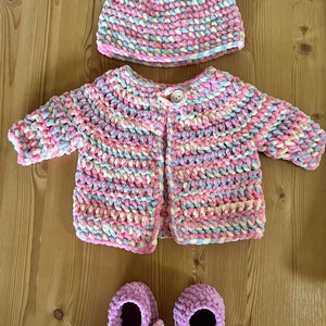 May include: A baby outfit set in pink, blue, and white. The set includes a crocheted cardigan, a crocheted hat with bear ears, and a pair of crocheted booties.