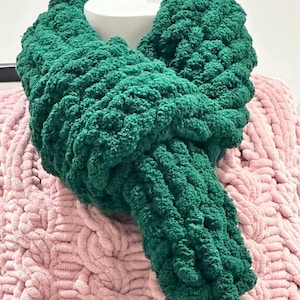 Può includere: Una sciarpa a infinito verde smeraldo, spessa, realizzata in filato morbido e strutturato. La sciarpa è drappeggiata su una forma cilindrica bianca e poggia su una coperta rosa a trecce. Il design ad anello della sciarpa suggerisce calore e comfort.