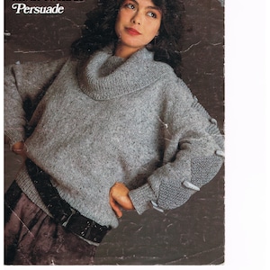 May include: A gray knitted sweater with a cowl neck and a textured pattern. The sweater has a loose fit and long sleeves. The sweater is shown on a model with a dark background.