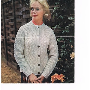 Puede incluir: Un cárdigan blanco de punto con un patrón de trenza y cierre de botones. El cárdigan lo lleva una mujer con el pelo rubio y está fotografiado contra una pared de ladrillo y vegetación.