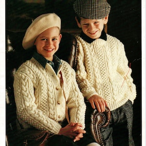 Puede incluir: Dos niños con suéteres de punto blanco con patrones de trenza. La niña lleva una boina blanca y el niño lleva una gorra de golf negra y blanca a cuadros. Los suéteres son ambos abotonados y tienen un diseño clásico de punto Aran.