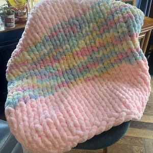 May include: A soft, hand-knitted blanket in pastel colors. The blanket features a large section of light pink at the bottom, transitioning to a section of pastel blue, yellow, and green. The blanket is made of thick, chunky yarn.