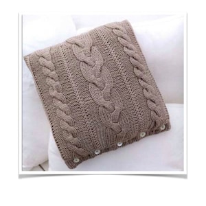 Peut inclure: Un coussin en tricot de câble brun avec des boutons blancs. Le coussin est sur une surface blanche.