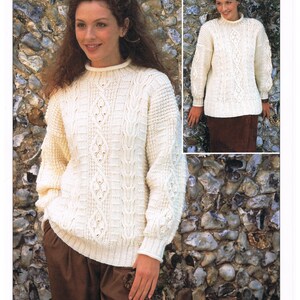 Puede incluir: Un suéter blanco de punto Aran con un patrón de punto de cable. El suéter tiene cuello alto y mangas largas. La modelo lleva pantalones marrones y está de pie frente a una pared de piedra.