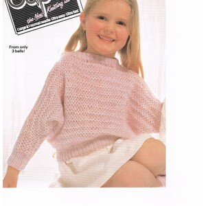 May include: A pink knitted jumper for a child, with a loose fit and a round neckline. The jumper is made with a brushed yarn and has a delicate openwork pattern. The jumper is shown on a young girl wearing a white skirt.