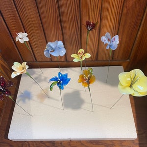 以下が含まれることがあります： 青、黄、赤、白など様々な色のガラスの花が11個、白い発泡スチロールの表面に並べられています