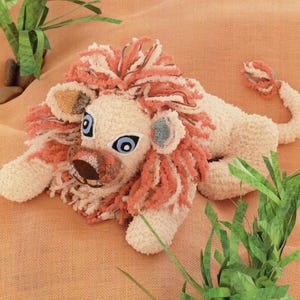 Op de afbeelding: Een gehaakt leeuwenknuffeltje met een wit lichaam, een roze manen en blauwe ogen. Het knuffeltje ligt op een bruine jute ondergrond met groen gras en stenen.