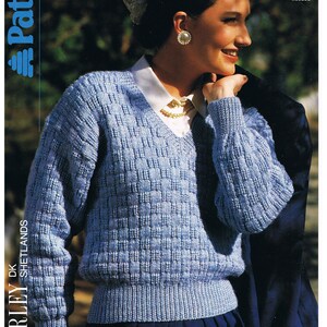 May include: A light blue knitted sweater with a v-neck and a textured pattern. The sweater is shown on a model wearing a white shirt and a dark blue skirt. The text "WAVERLEY SHETLANDS" and "Patons" is visible on the left side of the image. The text "PRICE CODE 3160 81-107 cm 32-42 in" is visible on the right side of the image. The text "Some experience needed" is also visible on the right side of the image.