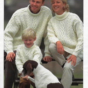 May include: A family of three wearing white cable knit jumpers with a dog. The jumpers have a traditional Aran design. The text "ARAN with 20% wool" is at the top of the image. The text "SIRDAR" is at the bottom of the image.