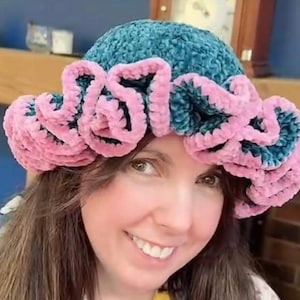 May include: A teal blue crocheted hat with a pink crocheted ruffle trim. The hat is worn by a person with long brown hair.