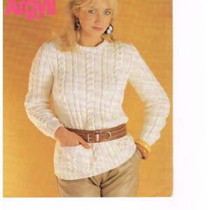 May include: A white cable knit sweater with a pink stripe detail. The sweater has a round neckline and long sleeves. The model is wearing a brown leather belt and a pair of brown pants.