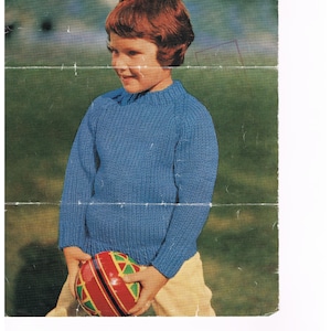 May include: A blue knitted sweater for children, with a round neckline and long sleeves. The sweater is being held by a child wearing beige pants. The image is from a vintage knitting pattern, with the text "Fleetknit Wool" and "6096 6d" visible.