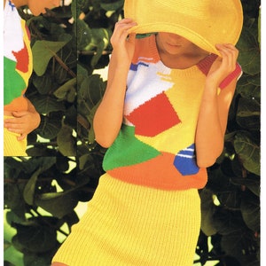 May include: A woman wearing a yellow knitted skirt and a colorful knitted vest with a geometric pattern. She is wearing a yellow sun hat and is standing in front of green foliage.