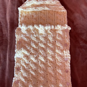 May include: A white knitted baby sleeping bag with a hood and bear-shaped buttons. The sleeping bag is made of a soft, fluffy yarn.