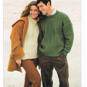 Puede incluir: Una pareja lleva suéteres, el hombre un suéter verde con un patrón de punto de cable y la mujer un suéter crema con un patrón de encaje. El hombre lleva pantalones de pana marrón y la mujer lleva pantalones marrones. La imagen tiene el texto "DOUBLE KNITTING" y "Hayfield".