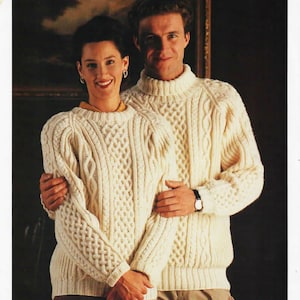 May include: A couple wearing matching white cable knit sweaters. The sweaters feature a traditional Aran stitch pattern. The man is wearing beige trousers and the woman is wearing a brown skirt.