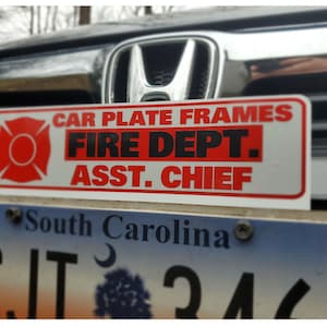 May include: A white car plate frame with red and black text that reads "CAR PLATE FRAMES FIRE DEPT. ASST. CHIEF". A red fire department symbol is on the left side of the frame. The frame is attached to a silver car with a South Carolina license plate.
