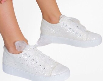 Könnte beinhalten: Ein Paar weiße Low-Top-Sneaker mit glitzernder Oberfläche, weißer Gummisohle und breiten, transparenten Organza-Schleifenbändern als Schnürsenkel. Die Sneaker verfügen über eine abgerundete Zehenkappe und silberfarbene Metallösen.
