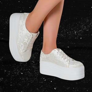 Puede incluir: Zapatillas blancas con plataforma y estampado floral. Los zapatos tienen suelas gruesas blancas y están atados con cordones. Las zapatillas están hechas de tela texturizada sobre un fondo negro.