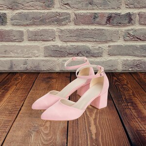 May include: A pair of light pink suede heeled shoes with ankle straps. The shoes have a pointed toe and a block heel.