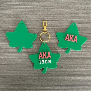May include: Green leaf-shaped keychains with the letters "AKA" in pink. One keychain has "1908" below the letters. The keychains have a gold-colored clasp and ring. The keychains are on a gray wooden surface.