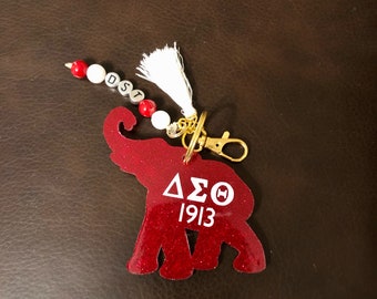 May include: Red glitter elephant keychain with a white tassel and a gold key ring. The elephant has the letters "ΔΣΘ 1913" printed on it.