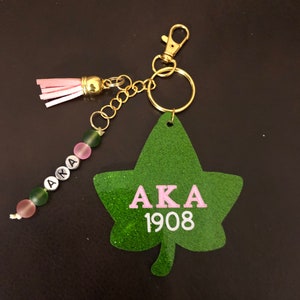 May include: Green glitter leaf-shaped keychain with pink letters spelling "AKA" and white numbers "1908". The keychain has a gold chain and a pink tassel.