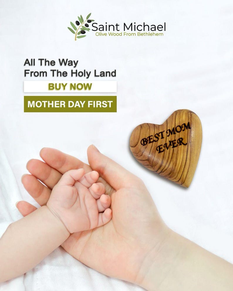 May include: A wooden heart-shaped ornament with the text "BEST MOM EVER" is held near a baby's hand in a larger hand. The image includes text that reads "Saint Michael Olive Wood From Bethlehem" and "All The Way From The Holy Land".