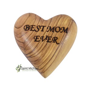 May include: A heart-shaped wooden carving with the words "BEST MOM EVER" etched in black. The olive wood has a natural grain pattern. The Saint Michael logo is in the lower left corner.
