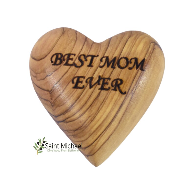 May include: A heart-shaped wooden carving with the words "BEST MOM EVER" etched in black. The olive wood has a natural grain pattern. The Saint Michael logo is in the lower left corner.