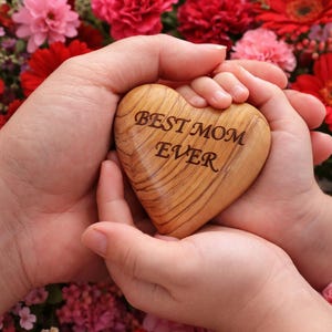 May include: A wooden heart with the inscription "BEST MOM EVER" is held in two sets of hands, surrounded by a vibrant array of red and pink flowers. The heart is light brown with visible wood grain.