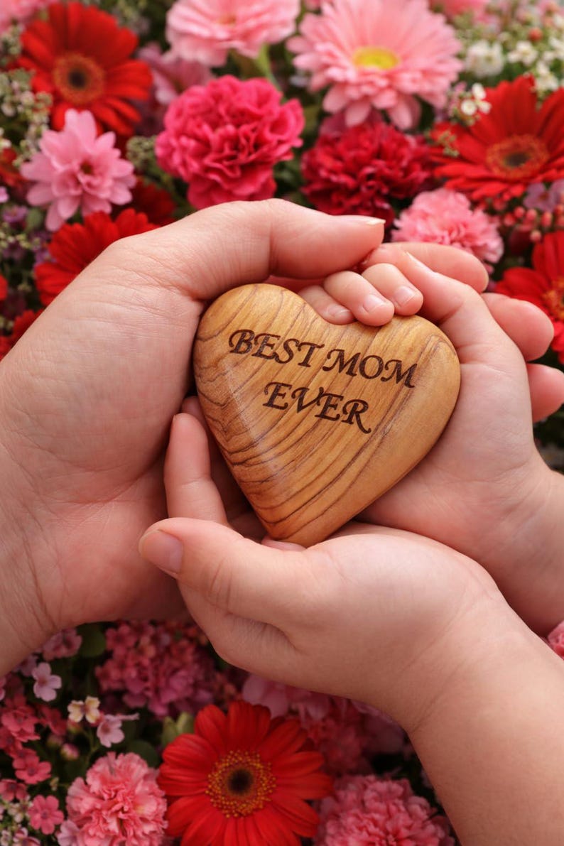 May include: A wooden heart with the inscription "BEST MOM EVER" is held in two sets of hands, surrounded by a vibrant array of red and pink flowers. The heart is light brown with visible wood grain.
