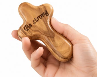May include: A wooden massage tool with a cross-like shape, held in a hand. The tool is made of light brown wood with visible grain patterns. The words "Be strong" are engraved on the surface.