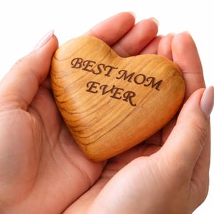 May include: A wooden heart-shaped keepsake with the inscription "BEST MOM EVER" in a dark brown font. The heart is held in open hands against a white background. The wood grain is visible.