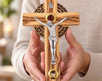 May include: A wooden crucifix featuring a silver-toned figure of Jesus, a circular backplate with the text GOD BLESS OUR HOME, a small gold-toned sunburst emblem above the figure, and a circular relic container at the bottom of the cross.