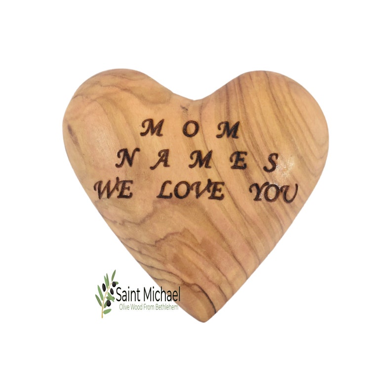 May include: Heart-shaped olive wood keepsake with the engraved text "MOM NAMES WE LOVE YOU." The wood has a natural grain pattern and a light brown color. The bottom left corner features the text "Saint Michael Olive Wood From Bethlehem."