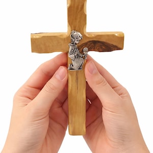 May include: A wooden cross with a silver-colored figure kneeling in prayer. The cross is made of light brown wood with a natural grain. The figure is holding a chalice and grapes. The cross is held by two hands.