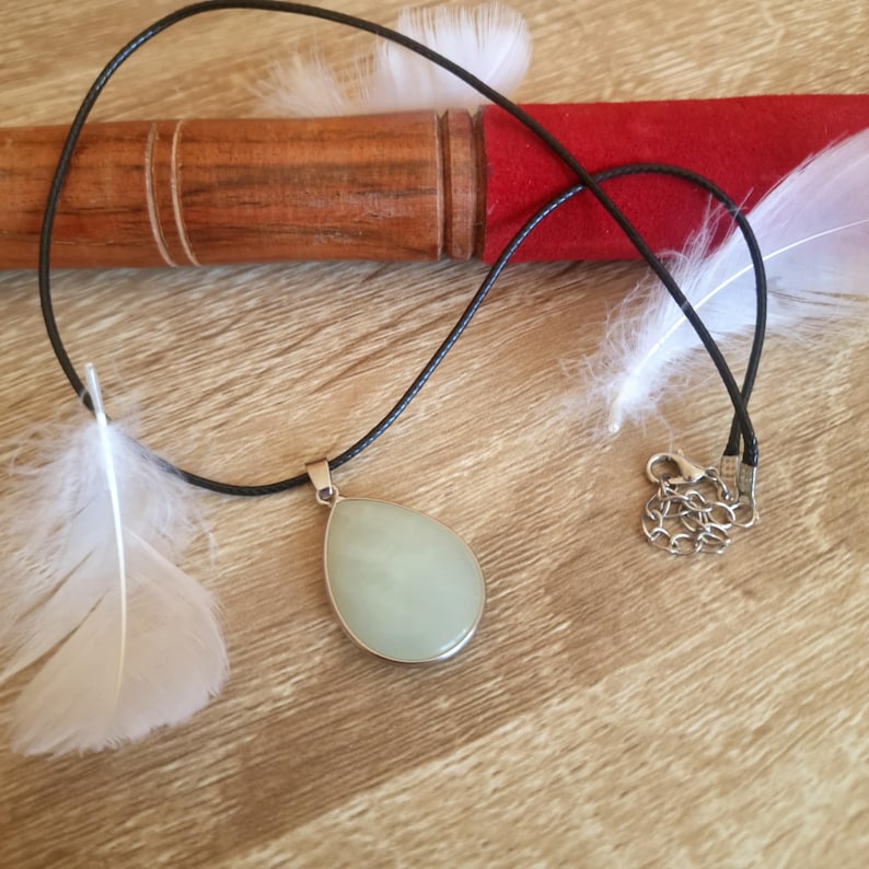 May include: A light green teardrop-shaped pendant necklace with a silver-colored bezel and a black cord. The necklace is displayed on a wooden surface with white feathers and a red and brown background.