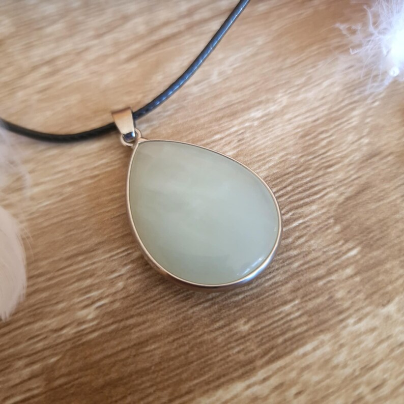 May include: A pear-shaped pendant with a pale green stone set in a silver-colored metal frame. The pendant hangs from a black cord necklace. The stone has a smooth, polished surface. The background is a wooden surface.