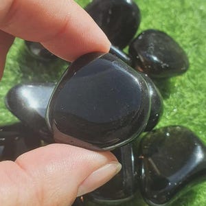Peut inclure: Plusieurs pierres d'obsidienne noires polies. Les pierres sont lisses, arrondies et réfléchissent la lumière, avec un aspect brillant. Les pierres sont présentées sur une surface verte.