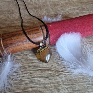 Peut inclure: Un collier avec un pendentif en forme de cœur en œil de tigre marron et doré. Le pendentif est suspendu à un cordon noir. Le collier est présenté sur une surface en bois avec un objet cylindrique rouge et des plumes blanches.