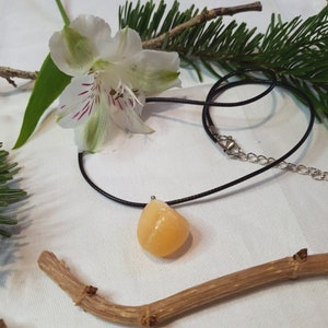 Peut inclure: Un collier pendentif avec une pierre précieuse en forme de goutte d'eau, de couleur orange clair. La pierre est suspendue à un cordon noir avec un fermoir argenté. Le collier est présenté sur une surface blanche avec des éléments décoratifs floraux et naturels.