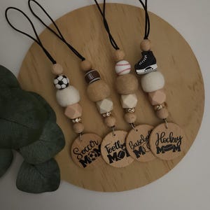 May include: Four wooden keychain charms with different sports themes. Each charm features a small wooden bead with a sports-themed image, a white felt bead, and a larger wooden bead with the text "Mom" and the sport name. The sports featured are soccer, football, baseball, and hockey.