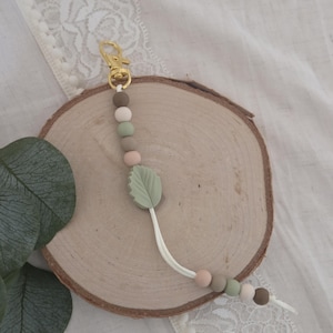 May include: A pacifier clip with a gold-colored clasp and a string of neutral-toned beads. A light green leaf-shaped charm is attached to the string. The clip is designed to attach to a pacifier or small toy.